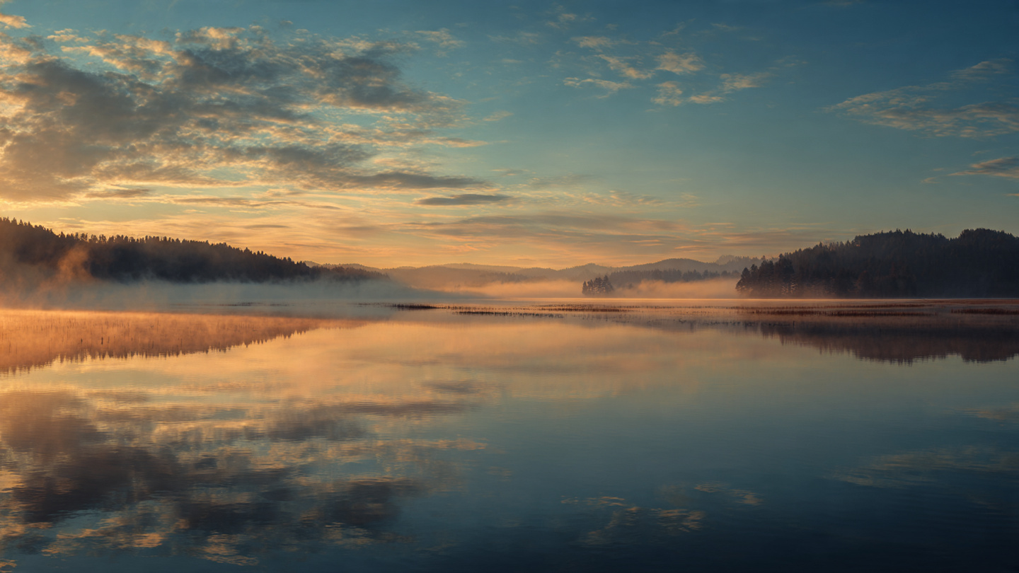 A sunrise over a calm lake with mist, creating natural patterns in the water and sky. Golden hour lighting, serene atmosphere