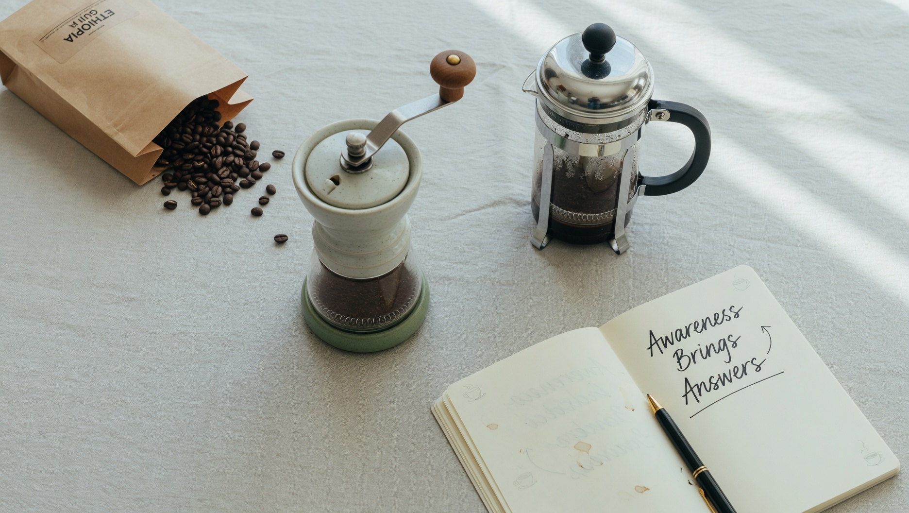 A minimalist flat-lay of coffee preparation tools: whole beans, ceramic grinder, French press, and a journal with 'Awareness Brings Answers' handwritten on the page, soft natural light, calming productivity aesthetic