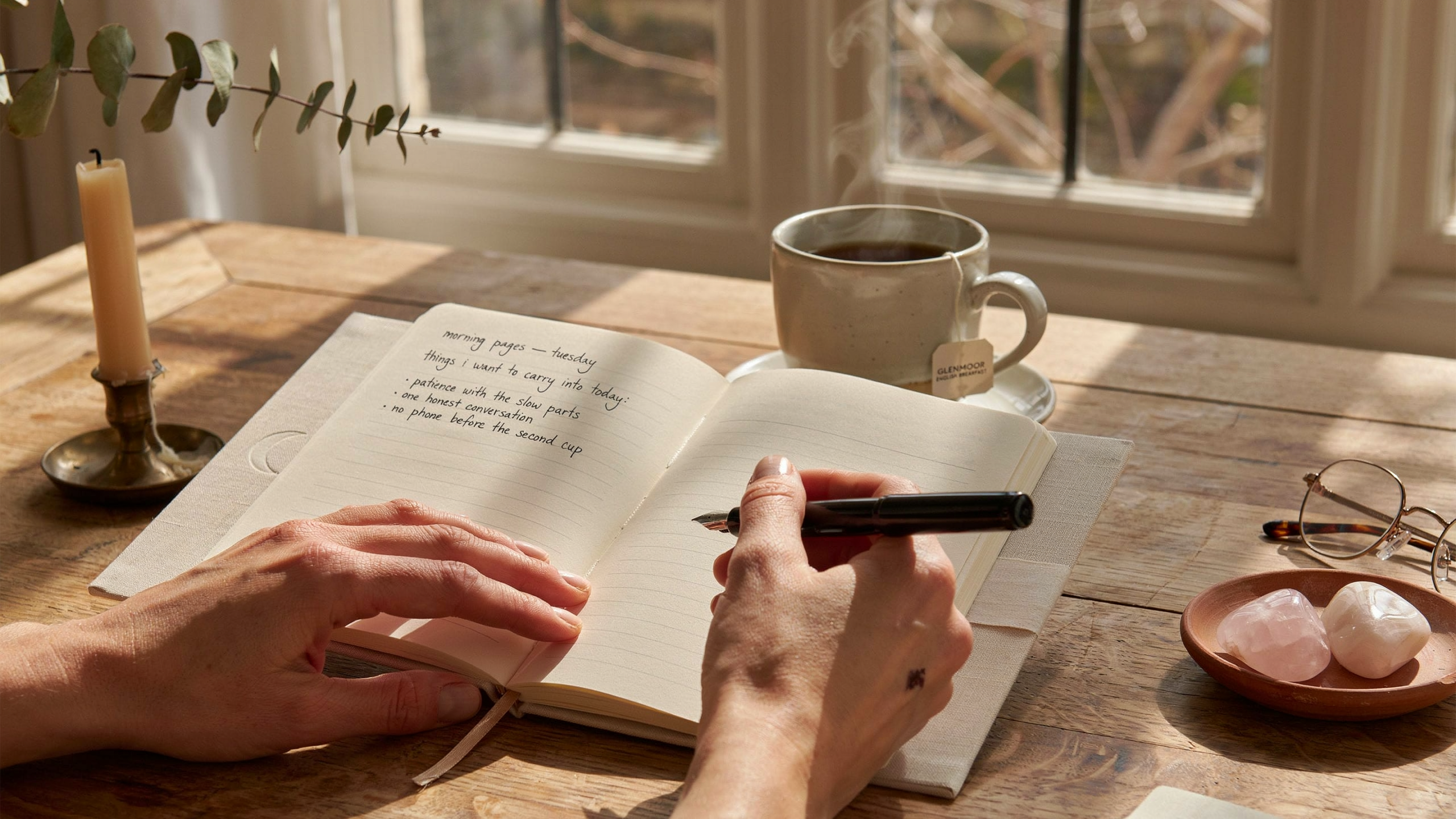 Close-up of hands holding a journal and pen, with a cup of tea nearby, morning light streaming through a window, symbolizing intentional morning practice and self-reflection. 