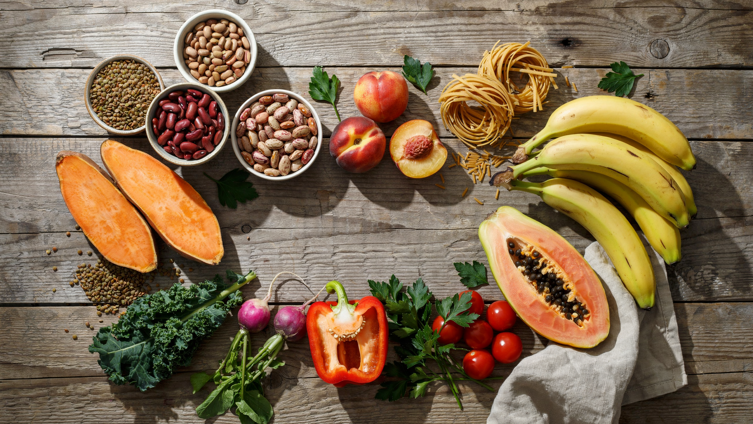 Photorealistic flat-lay photography of colorful, healthy foods: sweet potatoes, various legumes (lentils, kidney beans, pinto beans), fresh papaya, bananas, nectarines, whole grain pasta, and fresh vegetables. Arranged beautifully on a rustic wooden table with natural lighting. Photorealistic flat-lay photography of colorful, healthy foods: sweet potatoes, various legumes (lentils, kidney beans, pinto beans), fresh papaya, bananas, nectarines, whole grain pasta, and fresh vegetables. Arranged beautifully on a rustic wooden table with natural lighting.