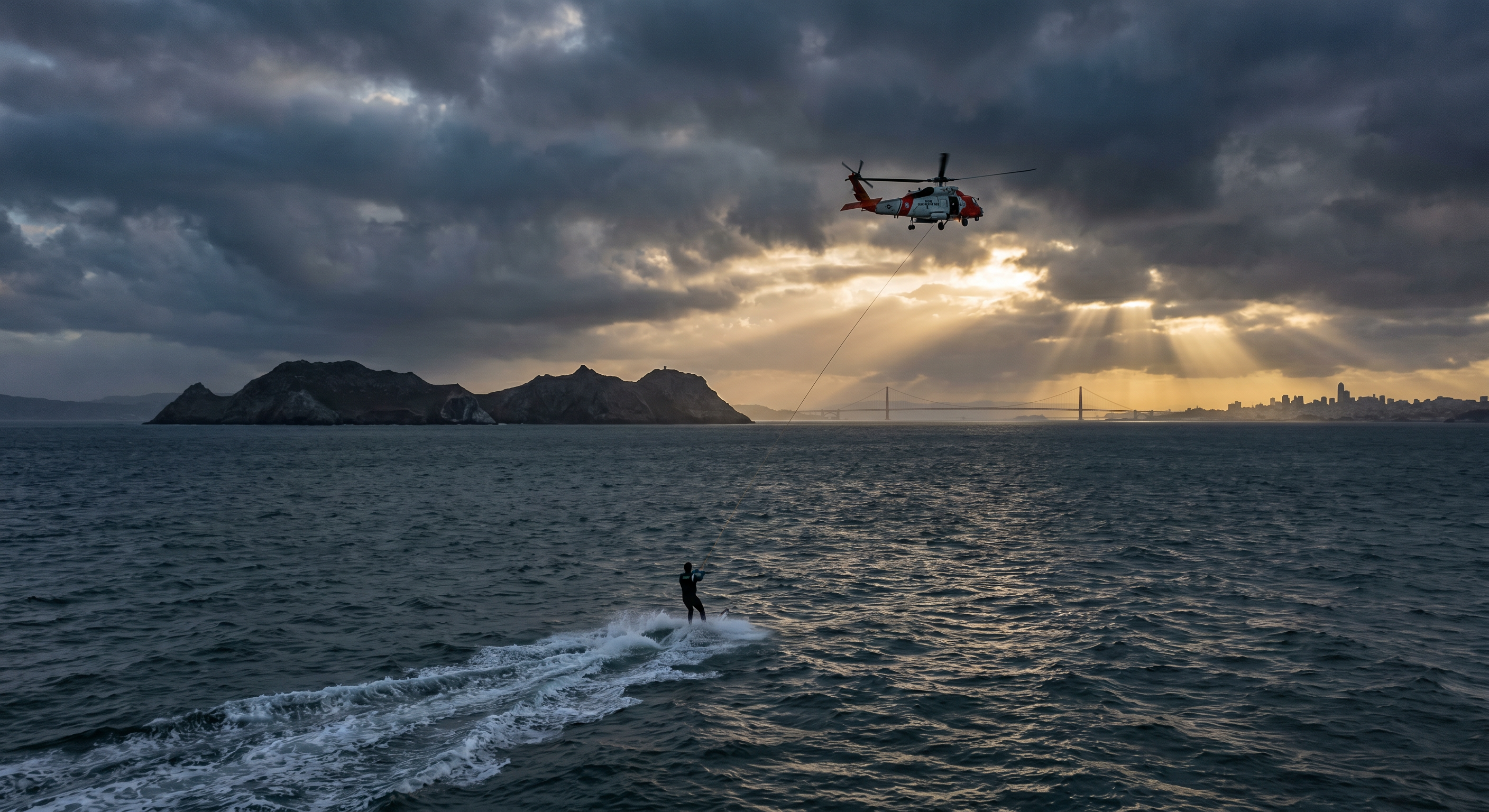 Dramatic aerial view of helicopter over San Francisco Bay with Farallon Islands in distance, stormy skies, water skier visible below on choppy water Being towed by the helicopter, There's a line, a tow rope, from the helicopter down to the skier on the water, as they are heading towards the island, away from the camera, tension and wisdom in the scene. Dramatic aerial view of helicopter over San Francisco Bay with Farallon Islands in distance, stormy skies, water skier visible below on choppy water Being towed by the helicopter, There's a line, a tow rope, from the helicopter down to the skier on the water, as they are heading towards the island, away from the camera, tension and wisdom in the scene.