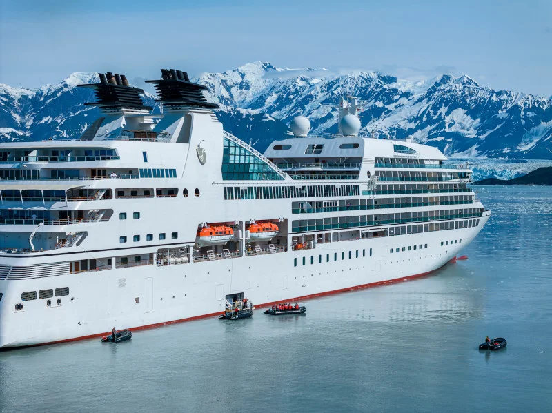 Small luxury cruise ship anchored near snow-capped mountains with tender boats ferrying guests to shore
