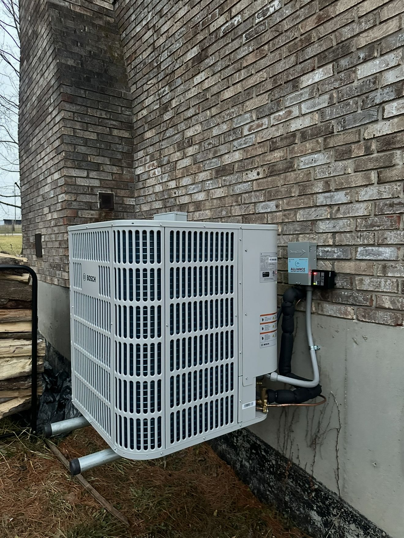 Bosch outdoor heat pump installed against a brick house, with an Alliance sticker on the electrical disconnect