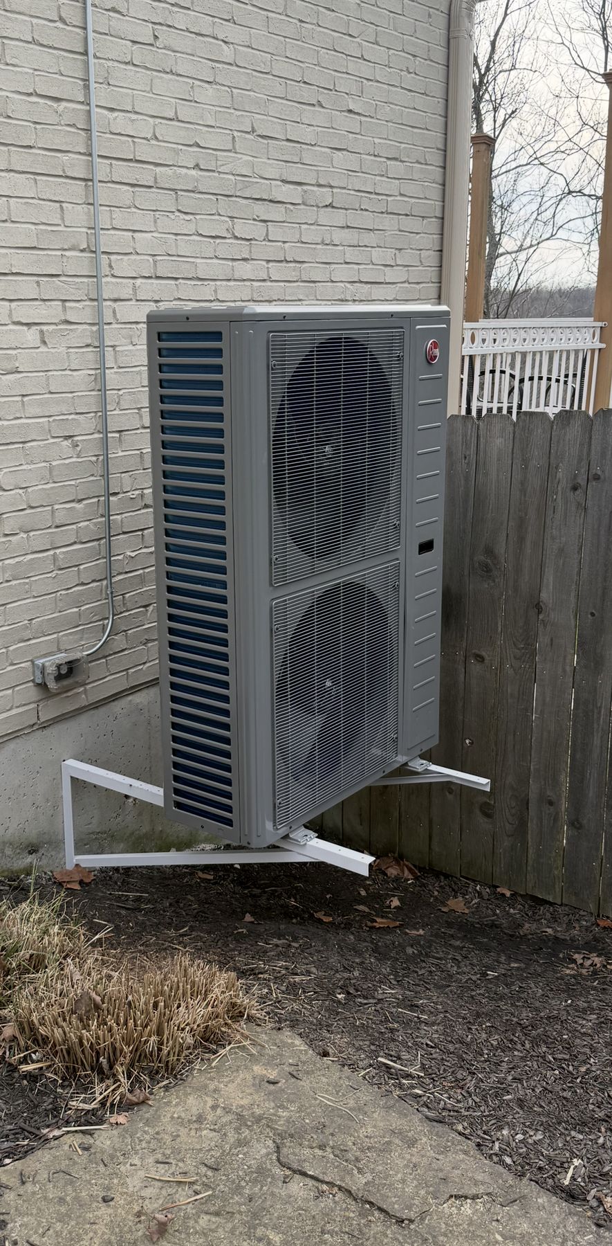 Rheem outdoor heat pump mounted on wall brackets against a white brick house