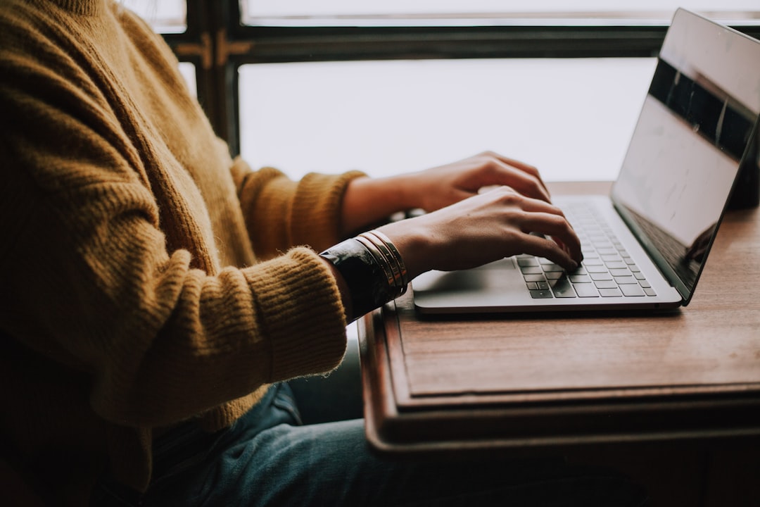 Cropped view of a person in a mustard knit sweater typing on a silver laptop at a wooden table beside a bright window.