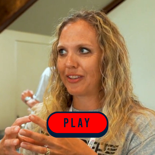A woman stands holding a sign that reads "Play," smiling and encouraging engagement in a playful activity.
