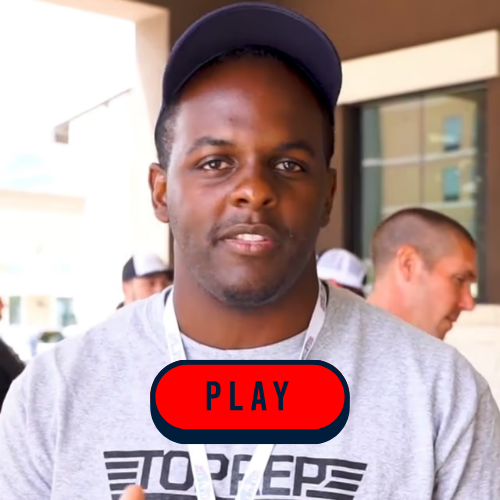 A man in a grey shirt holds a sign that reads "Play," smiling and standing in a casual outdoor setting.