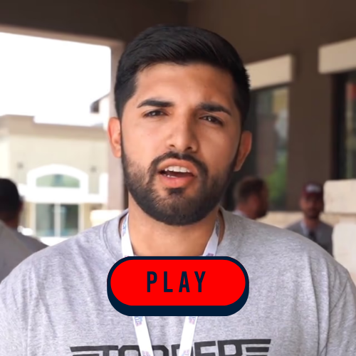 A bearded man in a grey shirt engages in conversation with another person.