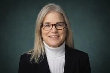 Studio headshot of Dr. Amy Taylor-Bianco in a white turtleneck and black blazer, against a dark teal background.