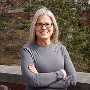 Dr. Amy Taylor-Bianco outdoors, smiling with her arms crossed, in a grey long-sleeved top, with evergreen trees behind her.