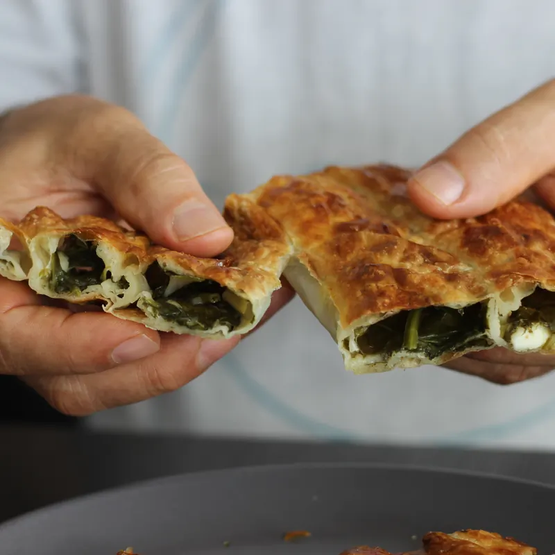 Hands tearing open a flaky spinach börek