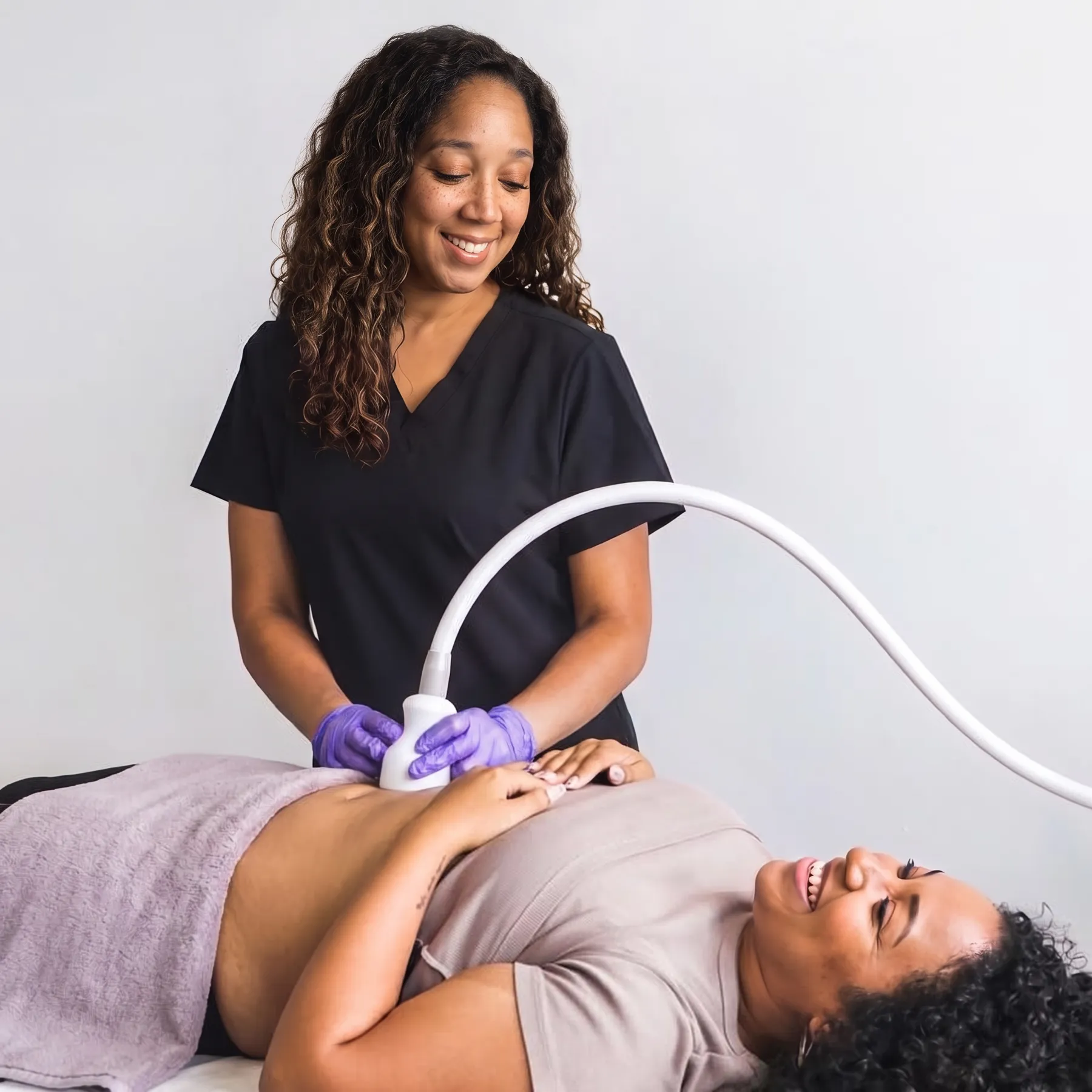 Specialist in black scrubs and purple gloves gliding a Cryoskin applicator over the abdomen of a smiling client lying on a treatment bed