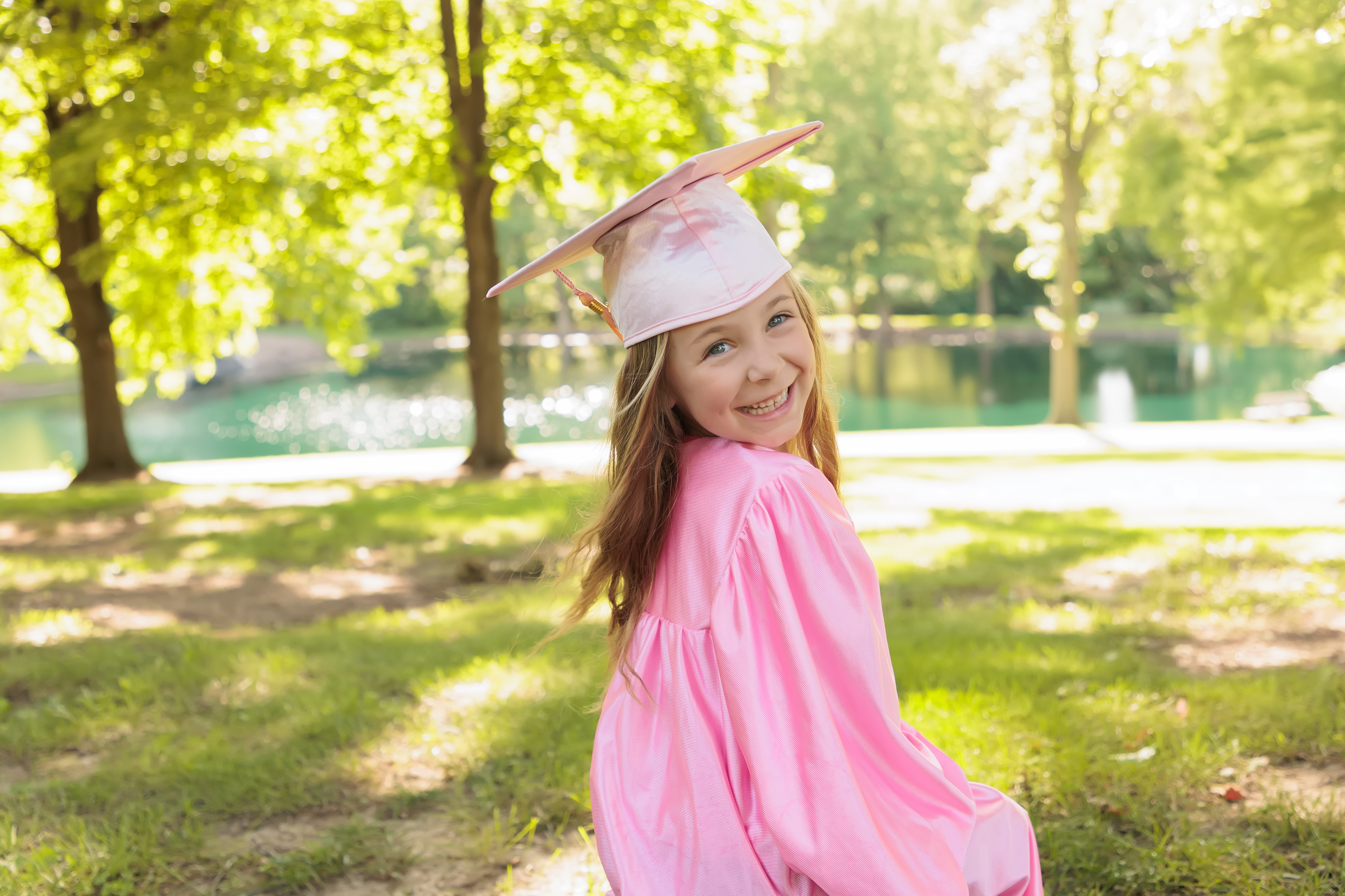 Little girl in a pink graduation cap and gown smiling over her shoulder in a sunny park