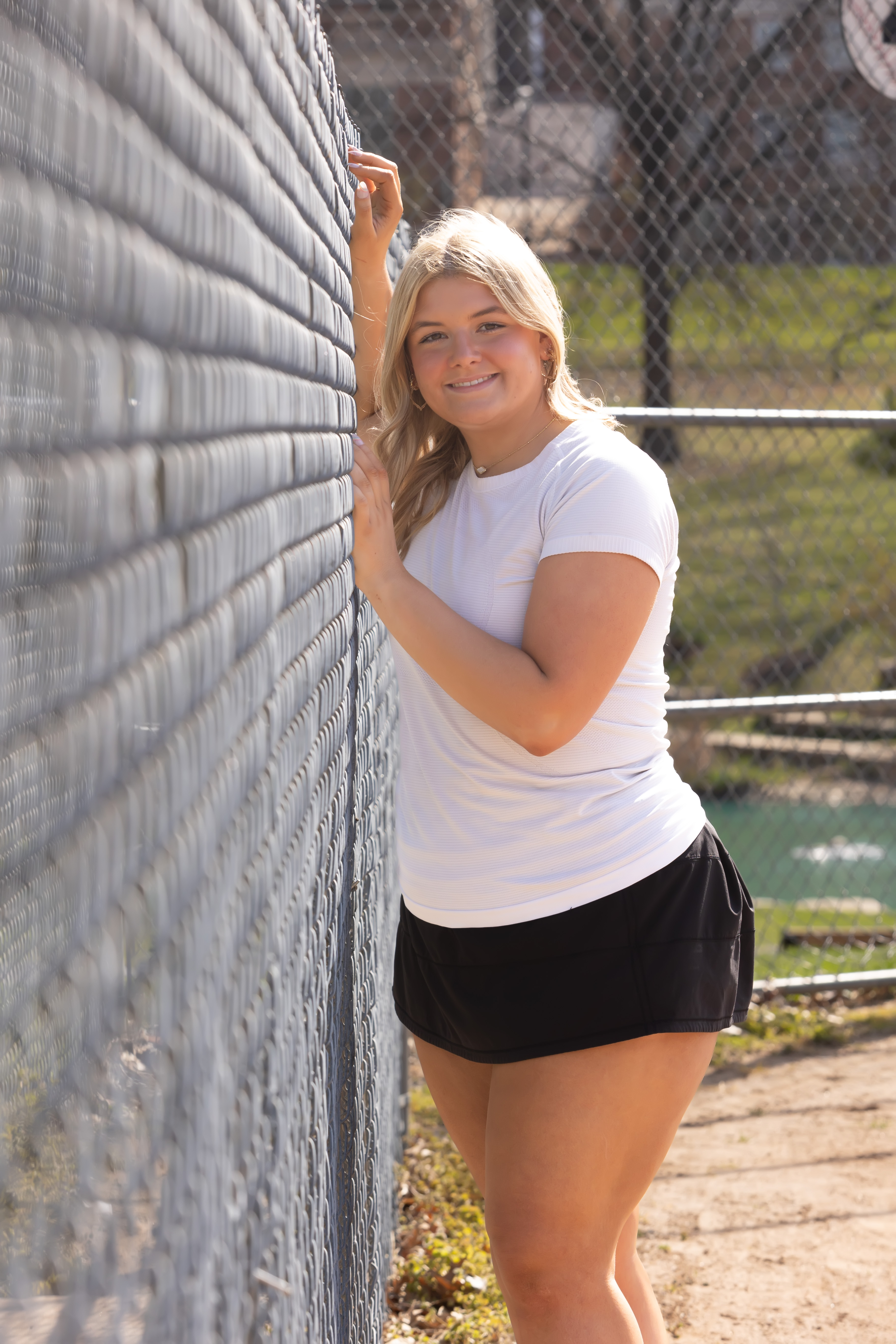 Senior girl in a white tee leaning against a chain-link ballfield fence