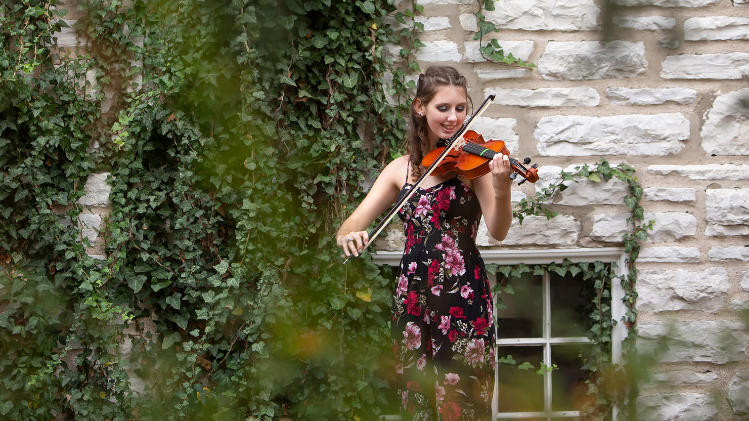 High school senior playing violin in a floral dress beside an ivy-covered stone wall