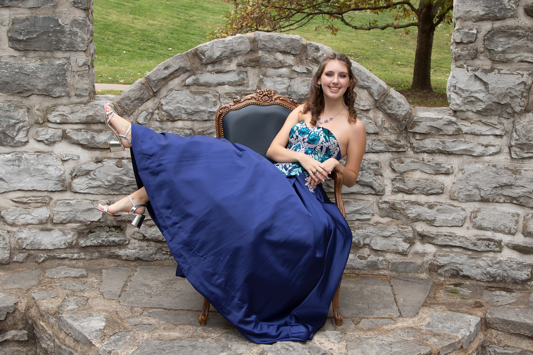 Senior girl in a navy ball gown lounging in an ornate chair in front of a stone wall