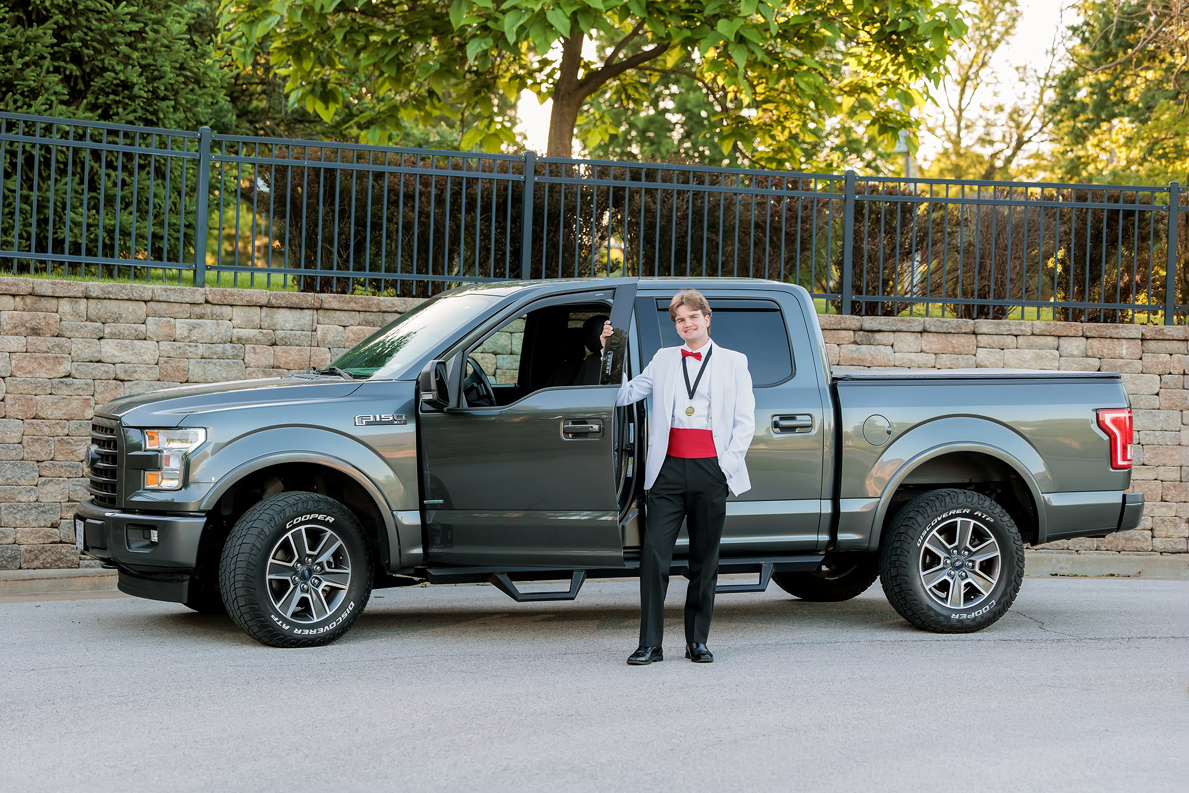 Senior in a white tux jacket and red cummerbund standing beside his gray pickup truck