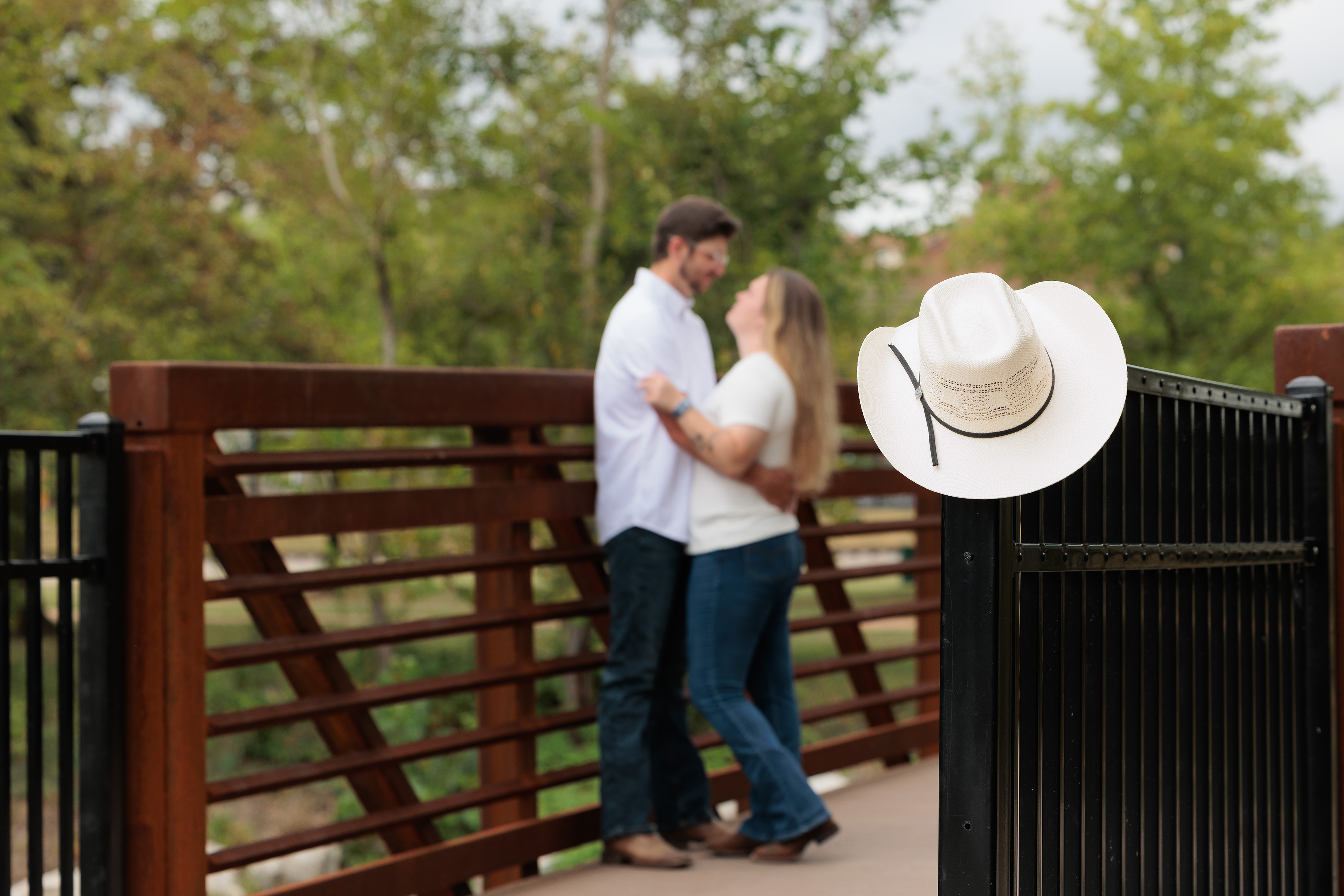 Couple embracing on a footbridge with a white cowboy hat on the railing, St. Louis engagement photos