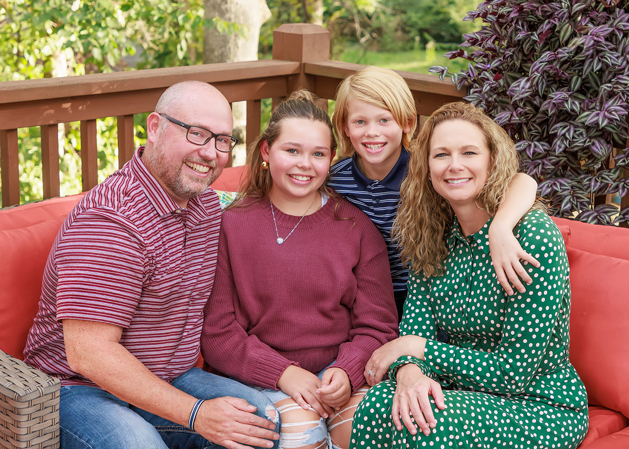 Family of four smiling together on a coral outdoor sofa on a backyard deck