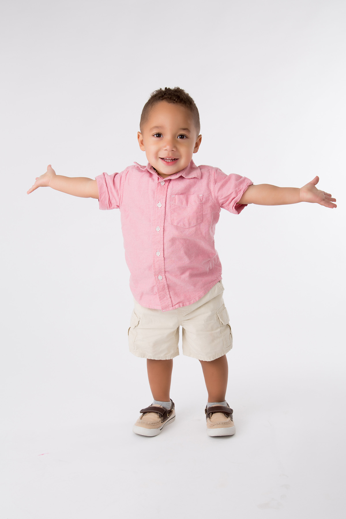 Smiling toddler boy in a pink shirt with his arms stretched wide, studio portrait