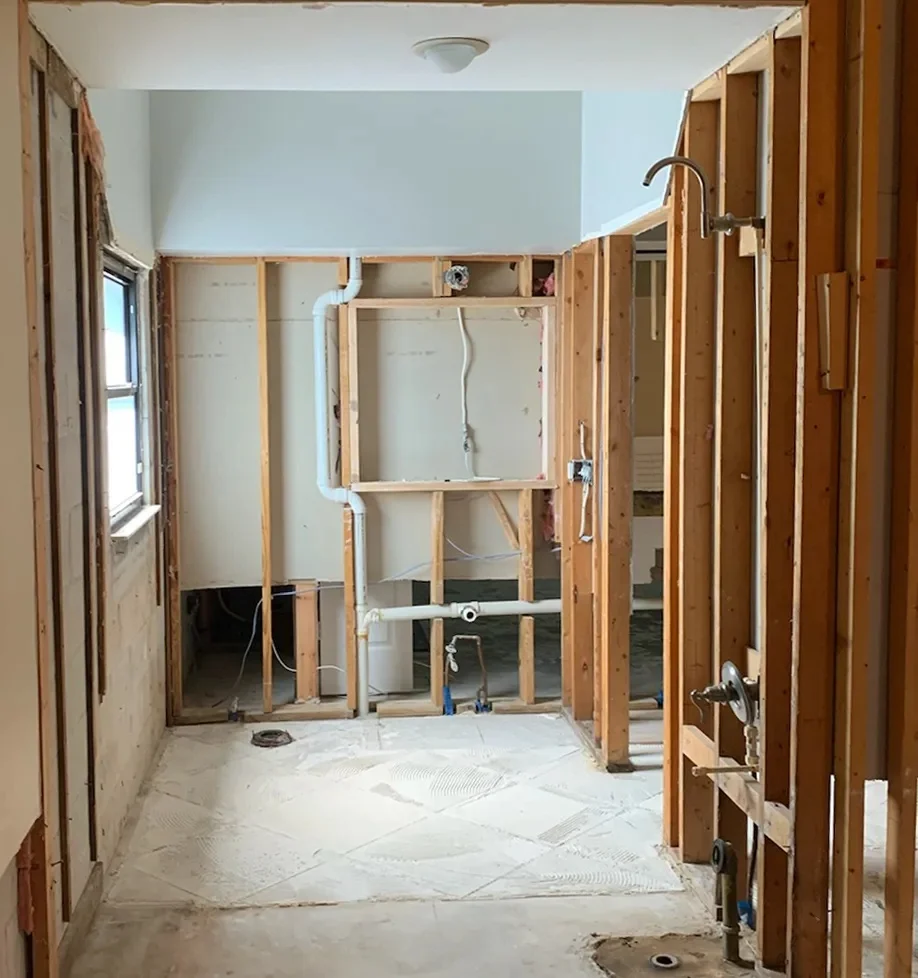 An unfinished bathroom shows wooden wall framing, exposed plumbing pipes, and a window, indicating a renovation in progress.