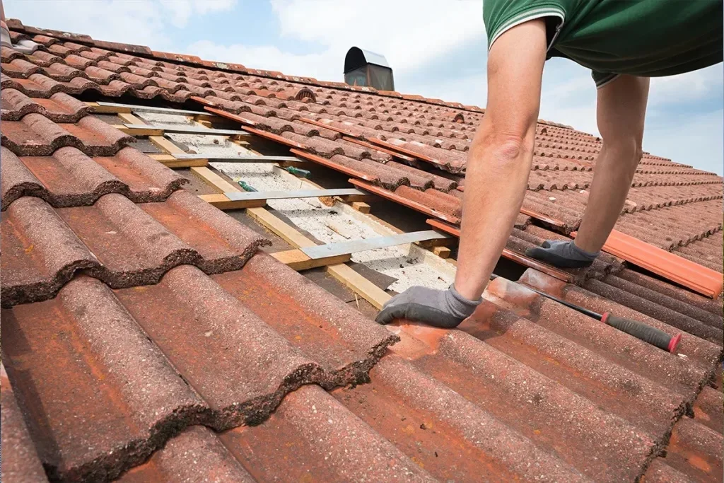 A person in gray gloves and green shorts works on repairing a tiled roof under a bright blue sky.