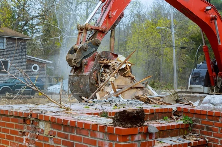 A red excavator's bucket filled with wood and debris from a demolished building, with a brick wall in the foreground.