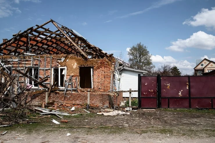 A severely damaged brick house with its roof completely destroyed and debris scattered on the ground under a cloudy sky.