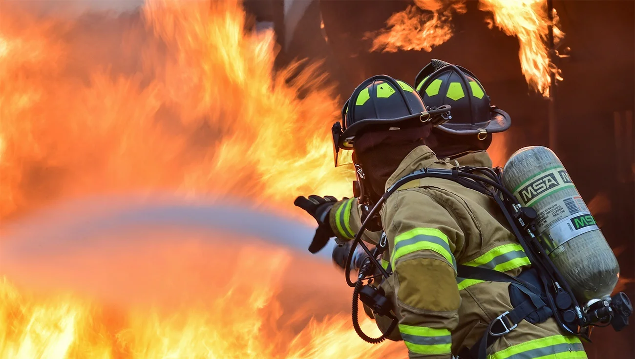Two firefighters in full gear spraying water into a large, intense blaze, seen from behind.