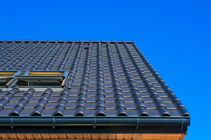 A close-up view of a dark tiled roof with two open skylights, a black gutter, and wooden eaves against a bright blue sky.