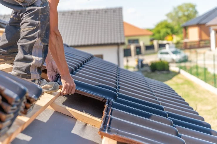 A close-up shot of a roofer's hands installing dark gray roof tiles on a wooden frame and underlayment.