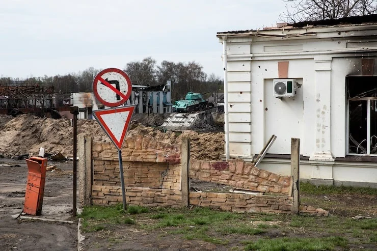 A war-damaged street features a bullet-ridden white building, fallen signs, and a tank on a dirt pile in the background.