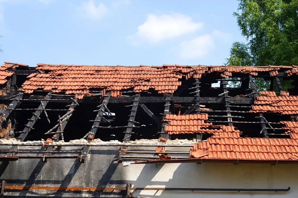 A house roof with significant damage, showing charred wooden beams and missing terracotta tiles against a blue sky.