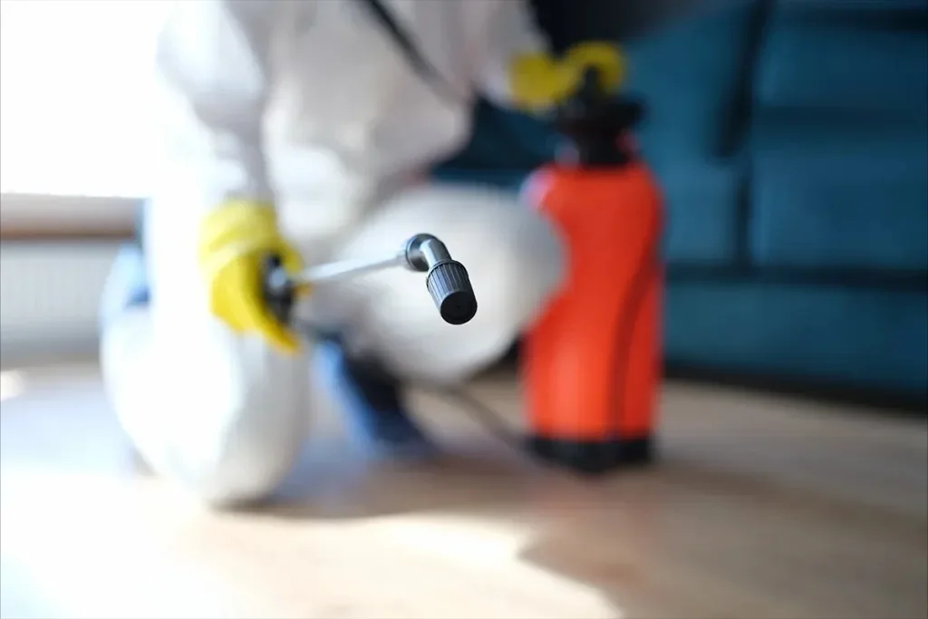 A close-up of a disinfectant sprayer nozzle on a light wooden floor, with a person in protective gear and an orange tank in the blurred background.