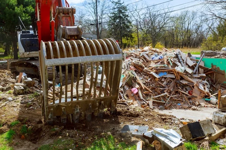A red excavator with a large claw bucket, positioned in front of a huge pile of wood debris and rubble from a demolished structure.