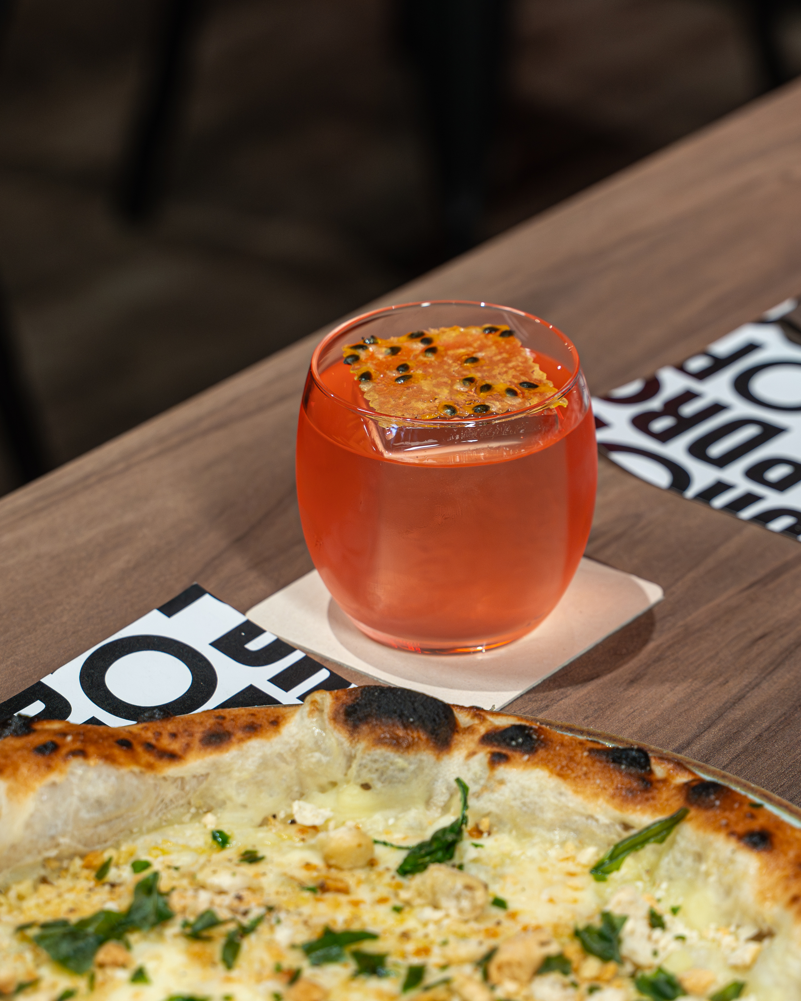 Un cocktail arancione con frutto della passione, servito accanto a una pizza DROP