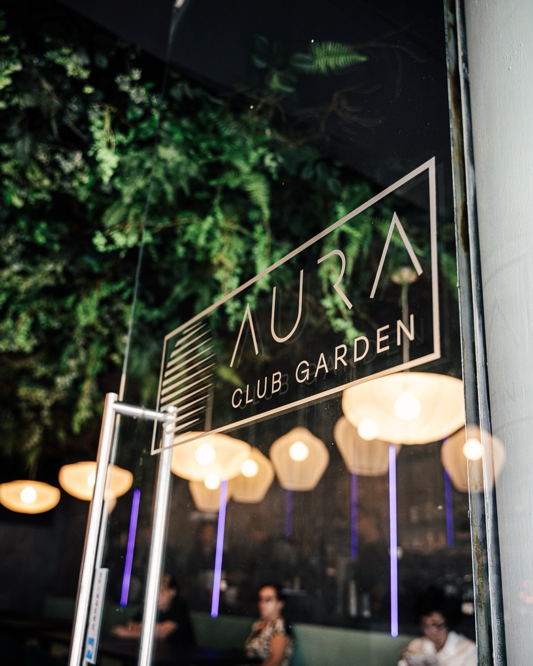 L'insegna AURA CLUB GARDEN illuminata tra le piante
