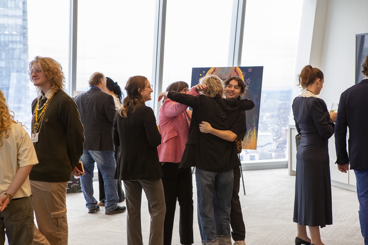 A group of people stand in a sunlit gallery with large windows overlooking a city, viewing artwork and talking, while two individuals share a hug in the centre during a Trans Day of Visibility art exhibit.