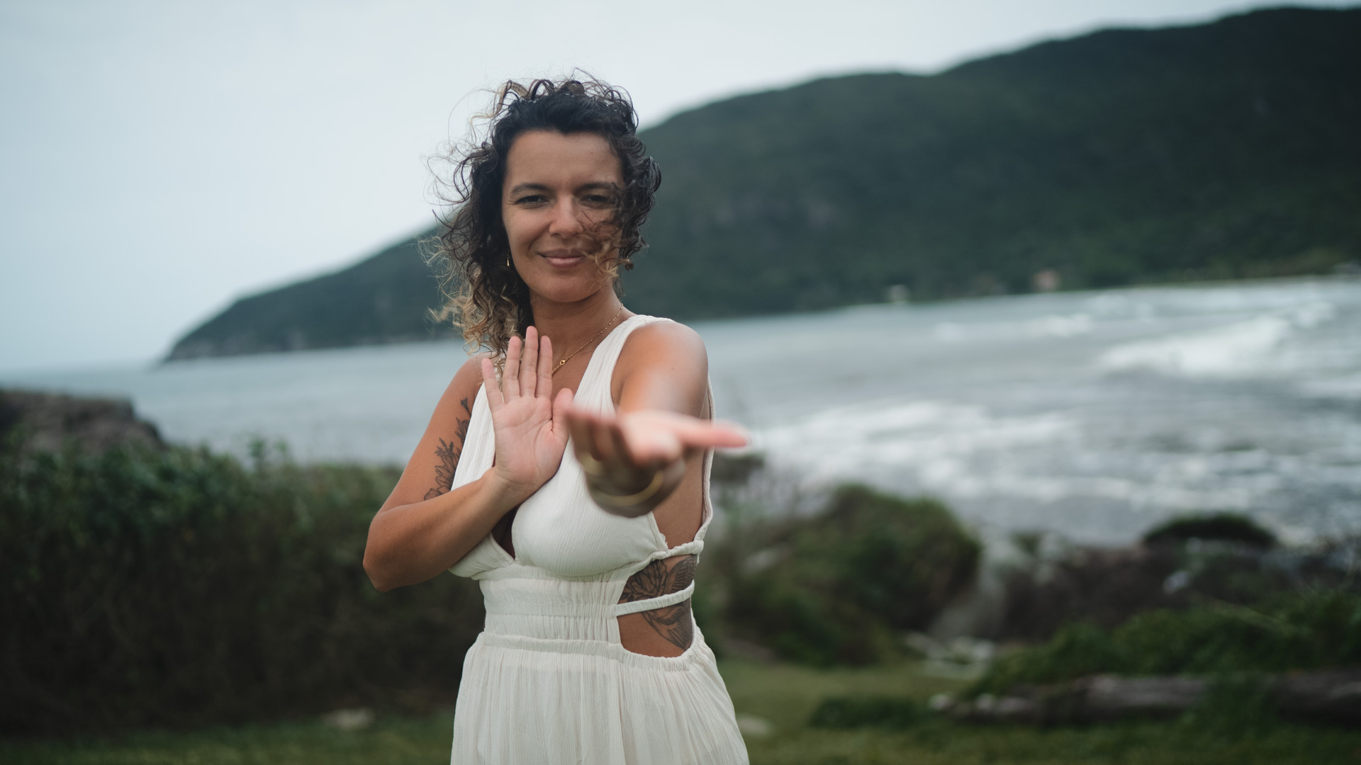 A woman in a white dress by the sea, reaching her open hands toward the viewer