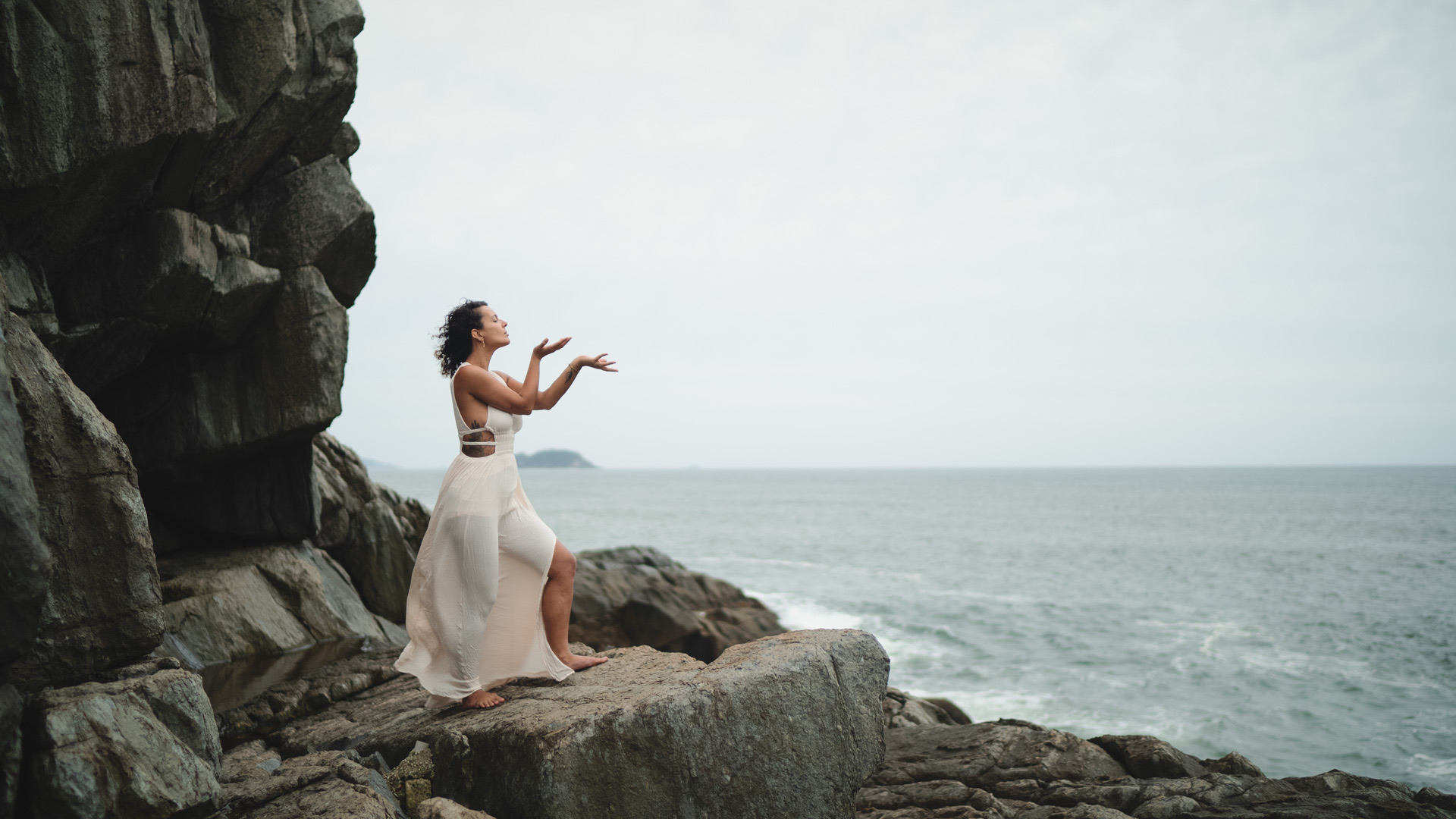 A woman in a white dress standing on coastal rocks, her hands raised toward the sky above the sea