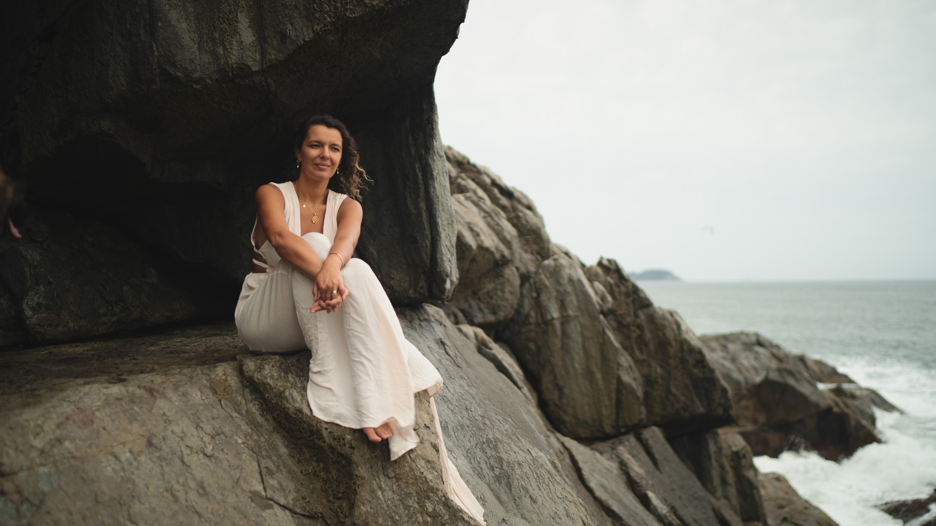 A woman in white seated on coastal rocks beneath an overhang, with the sea behind her