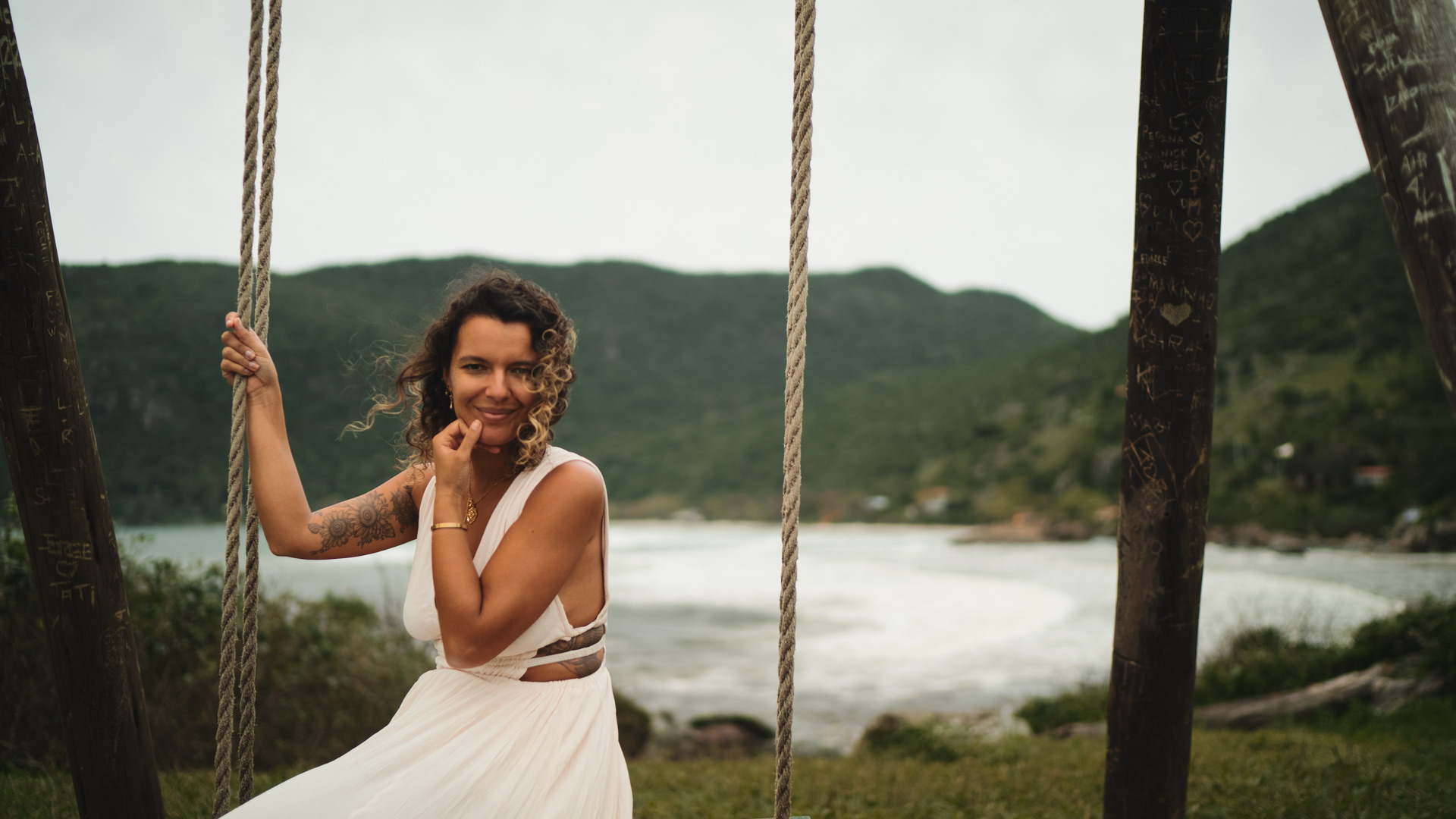 A woman in a white dress on a rope swing, smiling, with a bay and green hills behind her