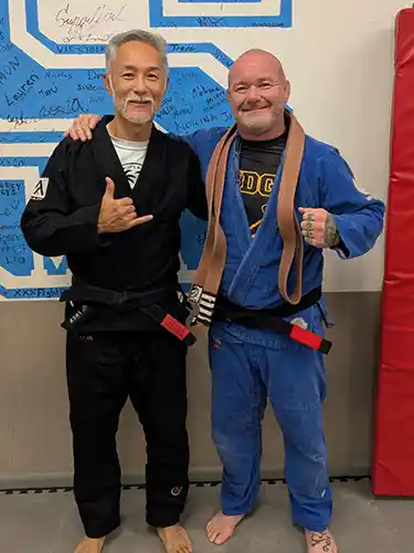 Two Jiu Jitsu coaches in their Gis at Stadium MMA in Port Orange, Florida, celebrating a belt promotion. The coach on the left is awarding a Black Belt to the coach on the right, and both are smiling proudly