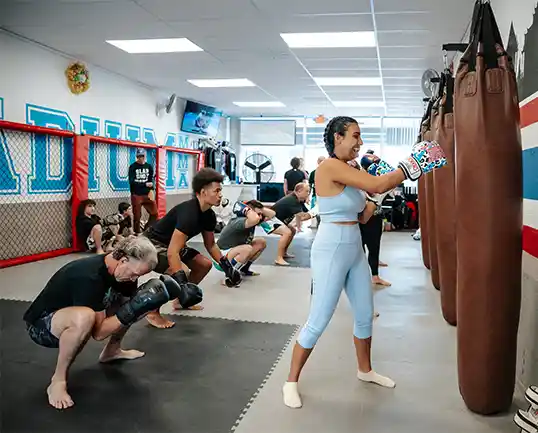 A diverse group participates in a kickboxing class at Stadium MMA in Port Orange, Florida, with half the students punching a heavy bag in boxing gloves and the others doing fitness exercises in the background