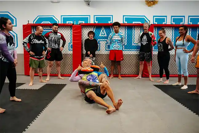 A diverse group of no-gi grapplers at Stadium MMA in Port Orange, Florida, watching their Jiu Jitsu coaches demonstrate a rear naked choke. The coaches have the class laughing, creating a fun and engaging training atmosphere