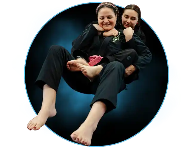 Two women in Brazilian Jiu Jitsu Gis practicing at Stadium MMA in Port Orange, Florida, with one applying a rear naked choke submission. Both are smiling and enjoying the training session