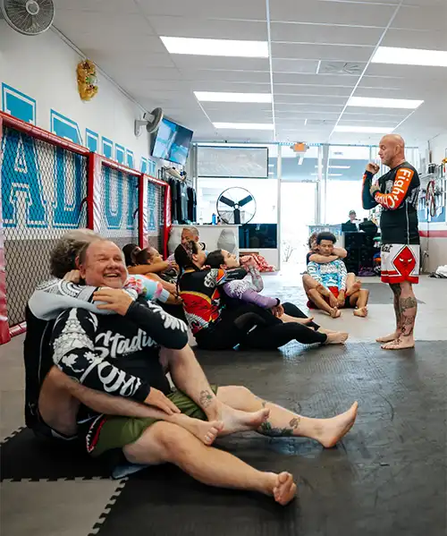 A diverse group of adults participating in a No Gi Jiu Jitsu class at Stadium MMA in Port Orange, Florida, with the instructor standing and providing guidance