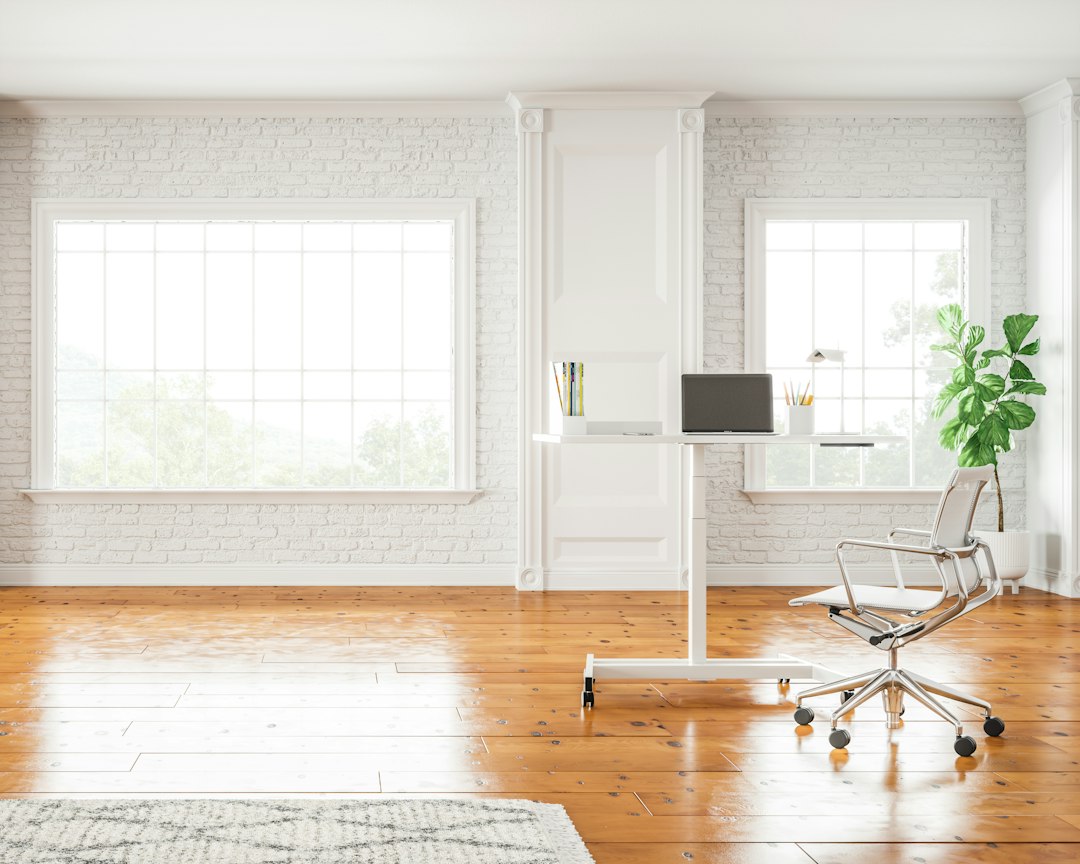 Sunlit room with white painted brick walls, large windows, wood floor and a small white standing desk with a laptop.