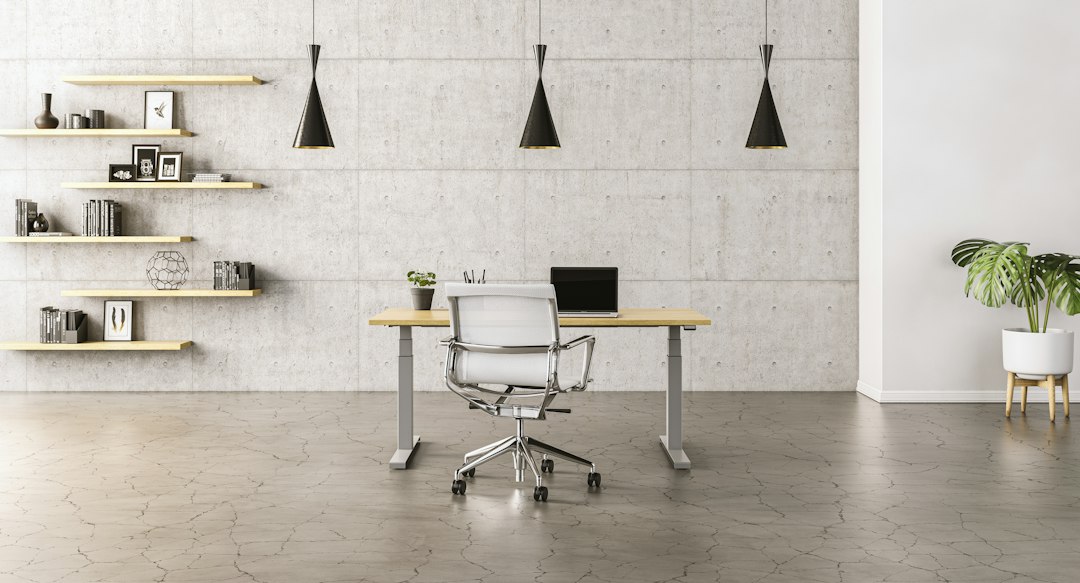 Minimal office with a height-adjustable desk, white mesh chair, floating shelves and black pendant lights against a concrete wall.