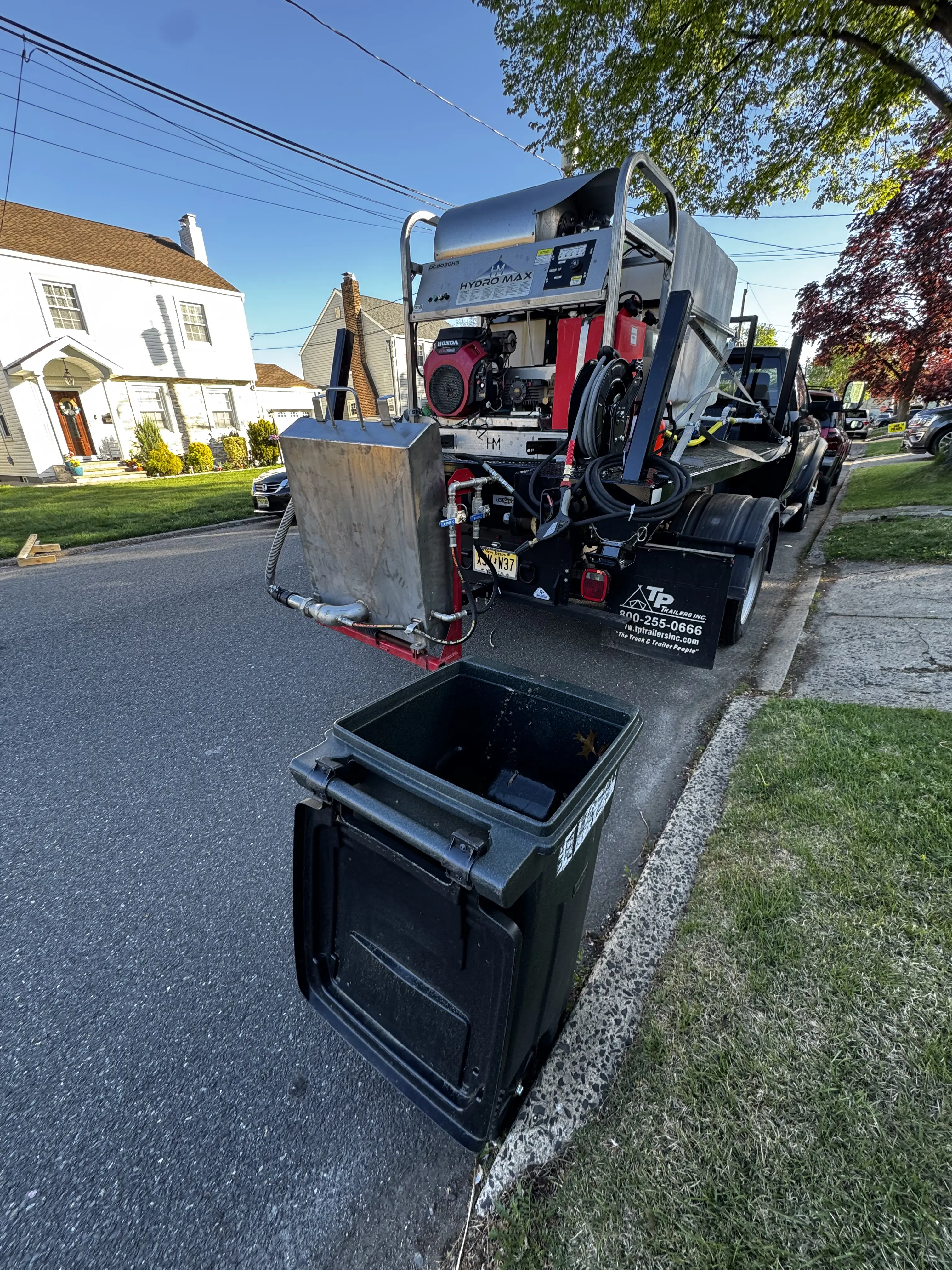 A black flatbed work truck parked on a brick driveway in a suburban neighborhood during sunny weather. The truck carries industrial equipment including a large water tank, hoses, and a pressure-washing or utility system mounted on the back. Trees, houses, and overhead power lines are visible in the background under a clear blue sky.